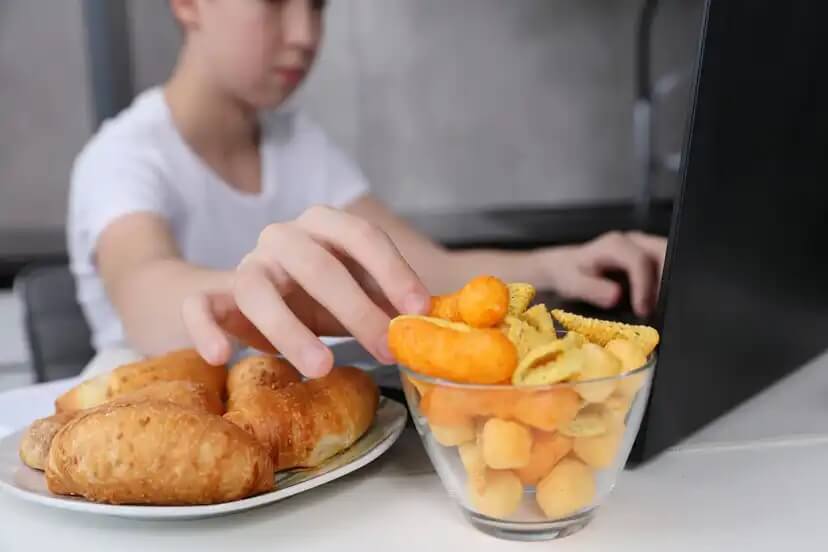 Criança comendo comida ultraprocessada enquanto joga no computador.